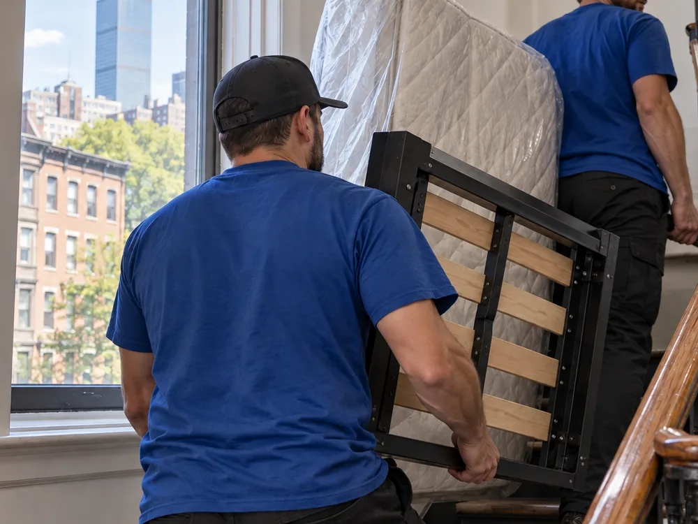 Movers carrying a disassembled bed frame and boxed mattress through a tight stairwell
